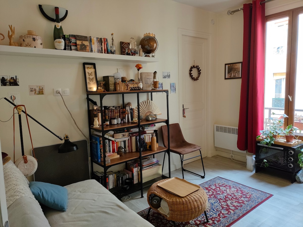 A cozy living area features a convertible sofa, a bookshelf filled with books and decorative items, and a chair. A round coffee table sits in front of the sofa. Natural light fills the space through large windows with red curtains, illuminating a small indoor plant.