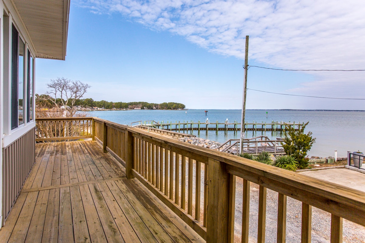 A spacious wooden deck overlooks a calm waterfront view, featuring distant trees and a well-maintained pier extending into the water. The clear sky and gentle clouds provide a serene backdrop, enhancing the outdoor space for relaxation and enjoying the surroundings.