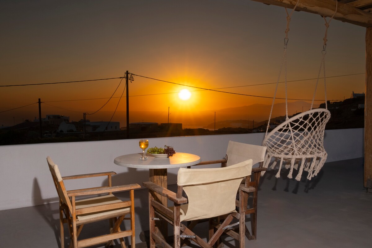 A private terrace features a round table and two wooden chairs, set against a stunning sunset backdrop. A hanging chair adds a relaxed touch, while a glass of beverage and a bunch of grapes sit on the table, enhancing the tranquil ambiance.