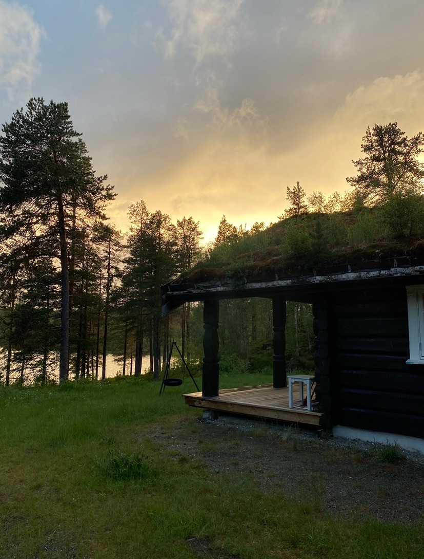 A charming timber cabin is set against a backdrop of tall trees and a lake at sunset. The overhanging balcony offers a serene view, while a grassy area surrounds the cabin. Soft lighting creates a tranquil atmosphere as the sun sets behind the foliage.