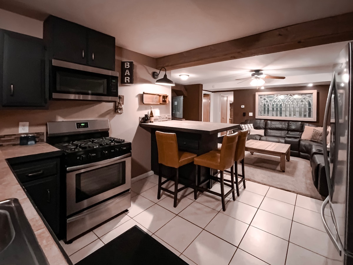 A functional kitchen area features stainless steel appliances, including a gas stove and a refrigerator. A dining table with two chairs is positioned near the kitchen, and a cozy living area with a sectional couch and coffee table is visible in the background.