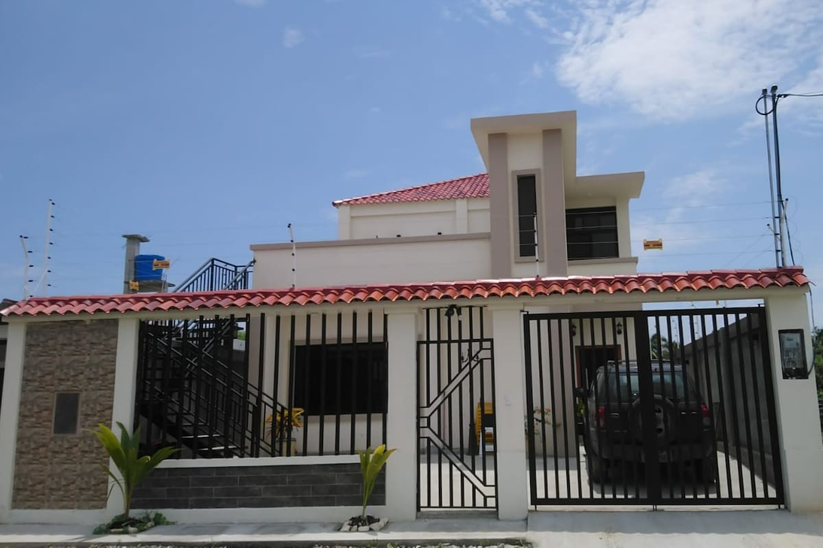 A modern two-story building features a red tiled roof and elegant exterior design. The entrance is marked by a gated front area with decorative plants. A staircase leads to a second-floor balcony, and windows allow natural light to brighten the facade.
