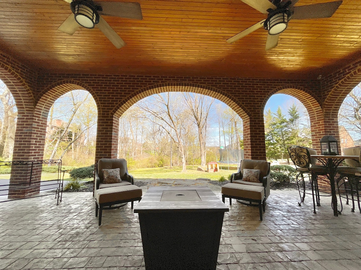 A covered outdoor porch features two comfortable chairs positioned around a low table. Arched brick supports frame the view of a lush backyard, showcasing trees and open green space. Subtle ceiling fans are visible above, enhancing the inviting ambiance.
