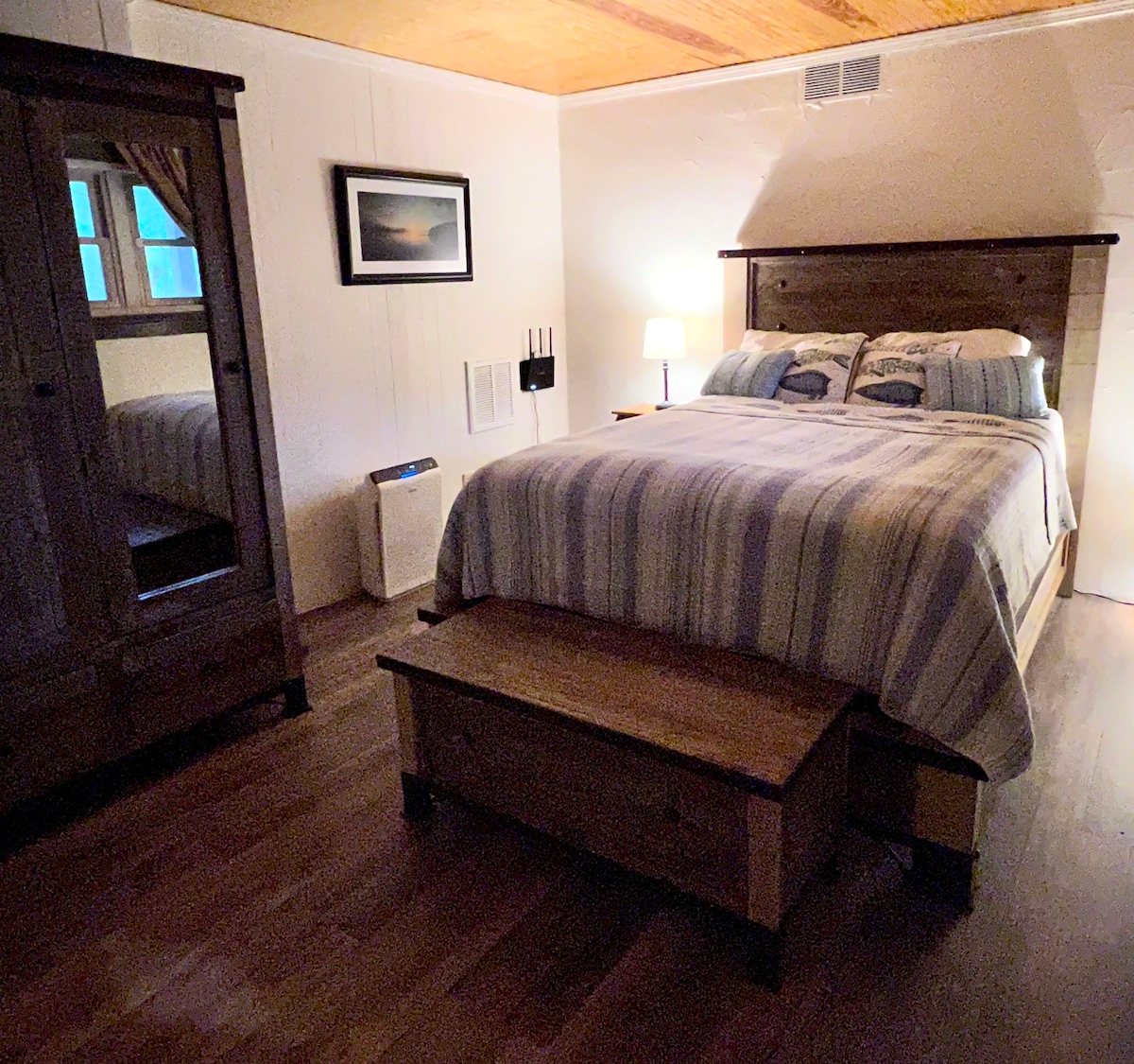 A cozy bedroom features a spacious bed with striped linens, complemented by a wooden bench at its foot. A light source beside the bed casts a soft glow, while a wardrobe with a mirrored door adds functionality. Light enters through a nearby window, creating a peaceful ambiance.