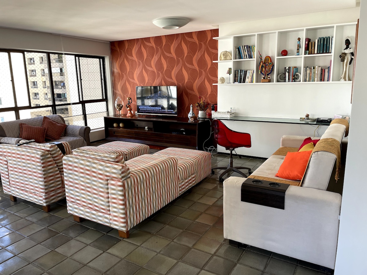 A spacious living area is furnished with a mix of patterned and solid-colored sofas, creating a comfortable environment. A flat-screen television is positioned on a low wooden unit, while a desk with a red chair is visible against a backdrop of bookshelves.