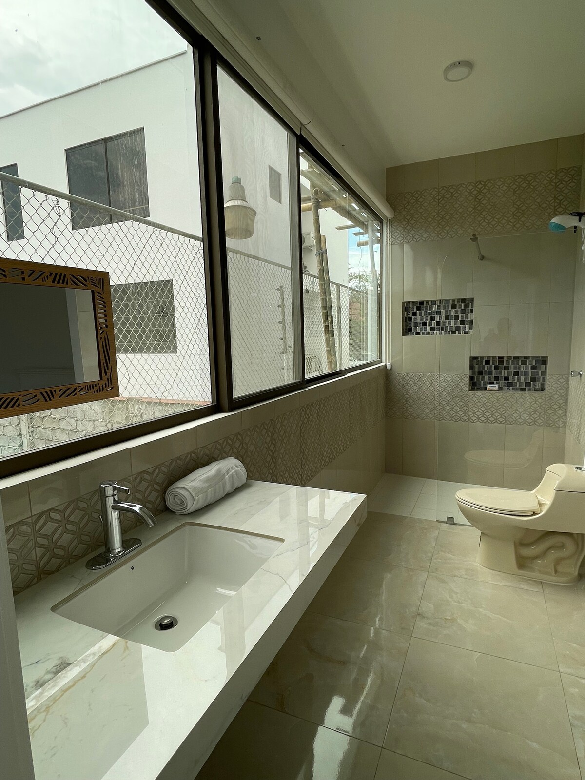 A spacious bathroom is featured with large windows allowing natural light. The vanity area includes a sink and is complemented by a large mirror. A modern toilet and a shower area with decorative tiles are also visible, creating a functional and clean environment.