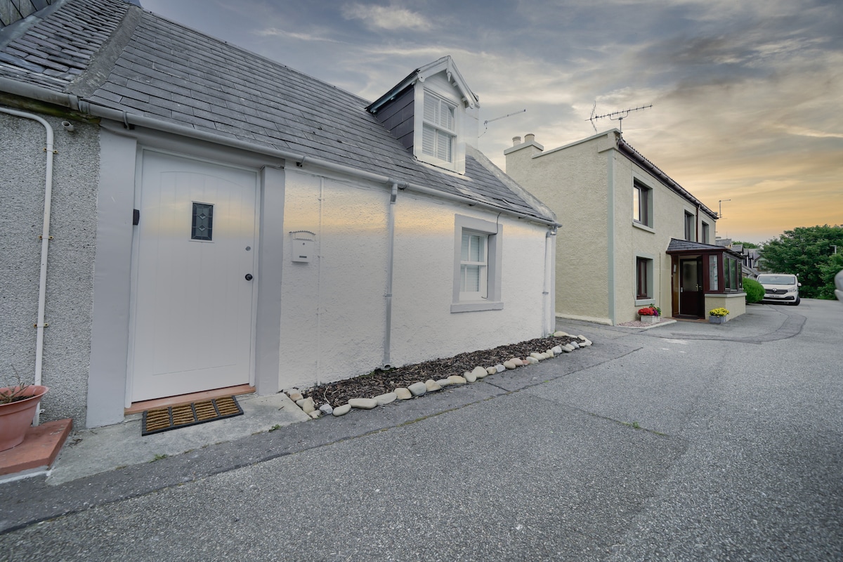 The charming exterior of the Wee House is shown, featuring a traditional grey stone façade with a welcoming white front door. A small garden area with stones borders the entrance, while neighboring houses can be seen lining the quiet street.