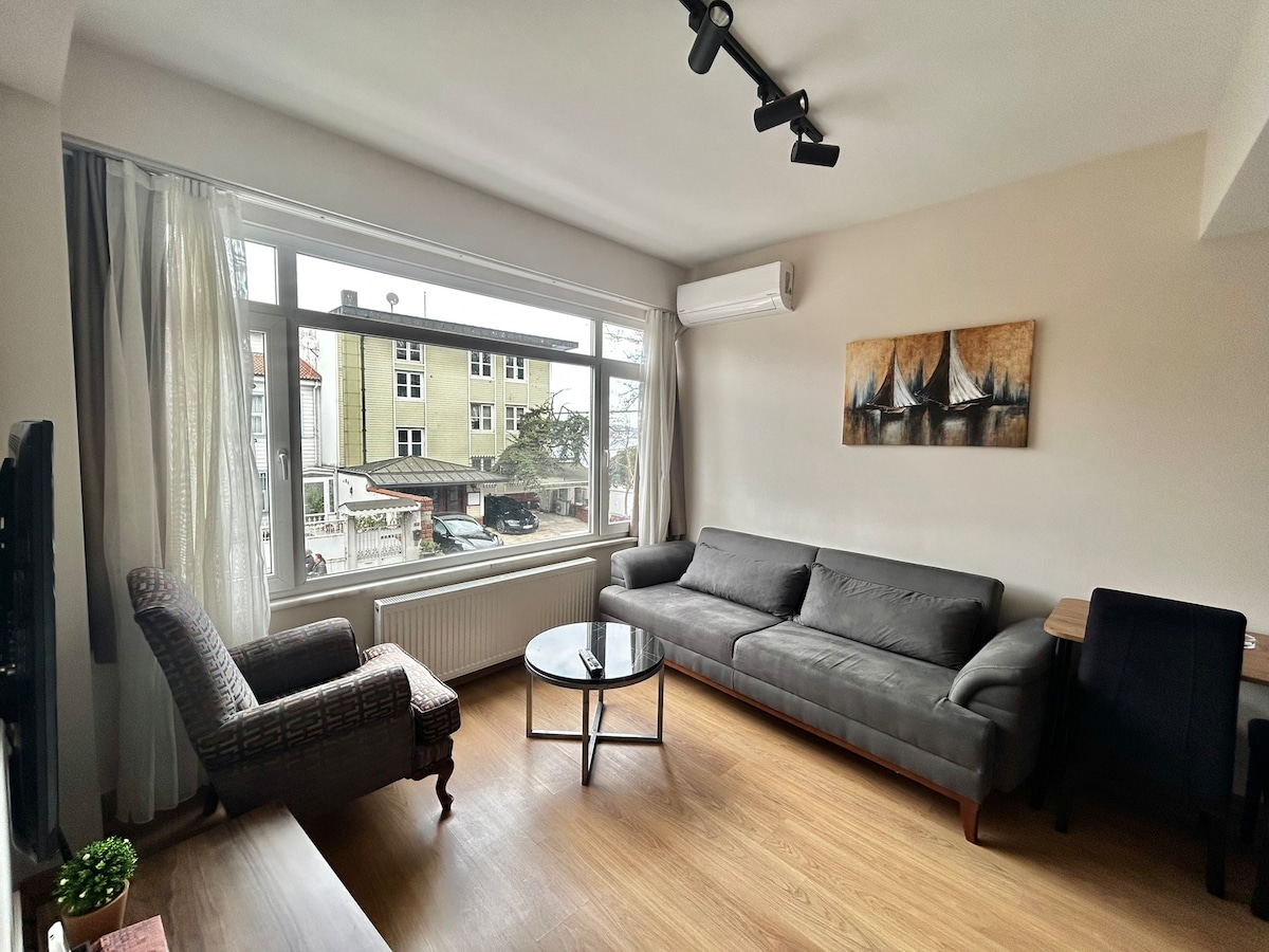A light-filled living area features a large window that allows natural light to enter. A gray sofa and an armchair are arranged around a circular glass coffee table. Neutral-toned curtains drape along the window, enhancing the space's calm atmosphere.
