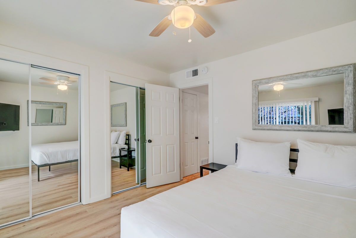 A light-filled bedroom features a spacious bed with white bedding. A mirrored closet reflects the room's design, and a ceiling fan is positioned above. A simple framed mirror adds depth, enhancing the room's inviting and airy feel.