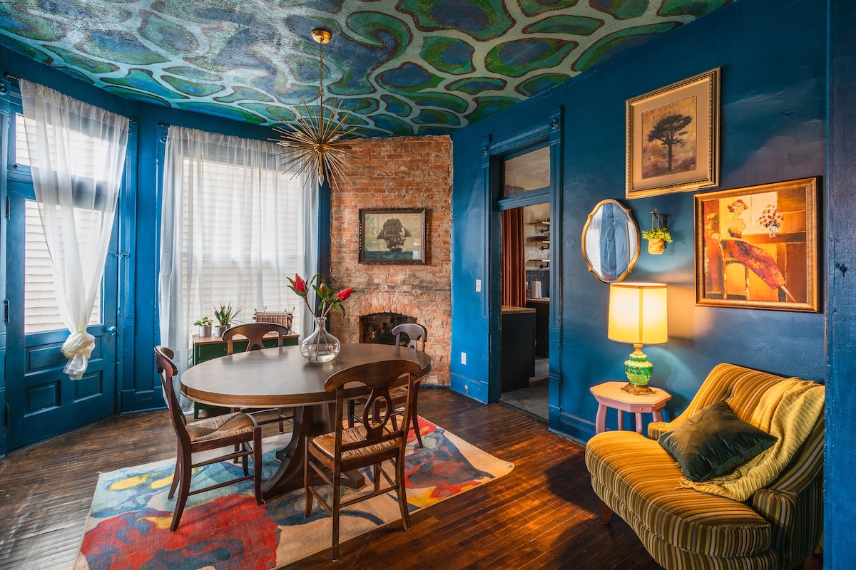 A dining area features a round wooden table surrounded by four chairs. The vibrant ceiling is painted with unique organic shapes, while the walls combine deep blue and exposed brick. A brightly colored rug adds warmth, and natural light filters through the sheer curtains.