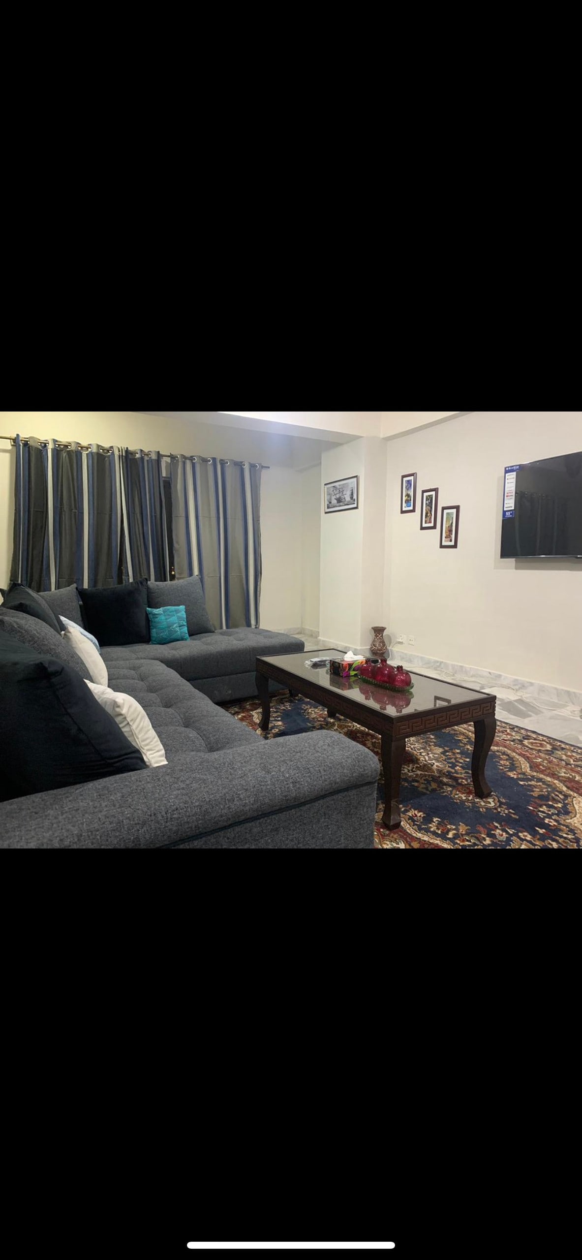 A cozy living area is furnished with a dark gray sofa adorned with several cushions in various colors. A wooden coffee table, decorated with a floral arrangement, is placed in front of the sofa. Wall-mounted framed pictures and patterned curtains contribute to the inviting ambiance.