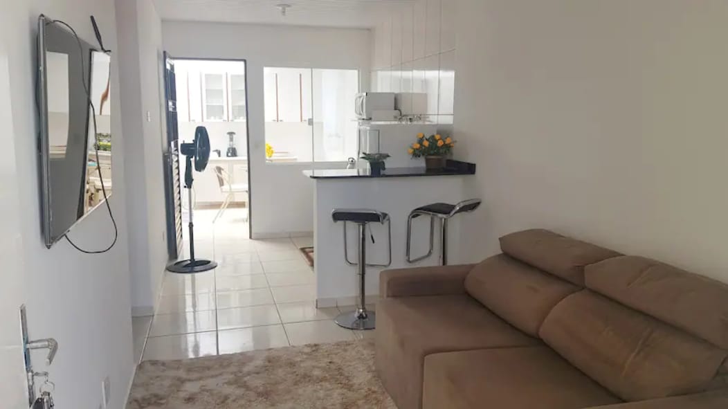 A light-filled living area features a cozy sofa, with a soft rug underneath. Bar stools are positioned at a kitchen counter visible in the background, where essential kitchen appliances are placed. A television hangs on the wall opposite the seating area.