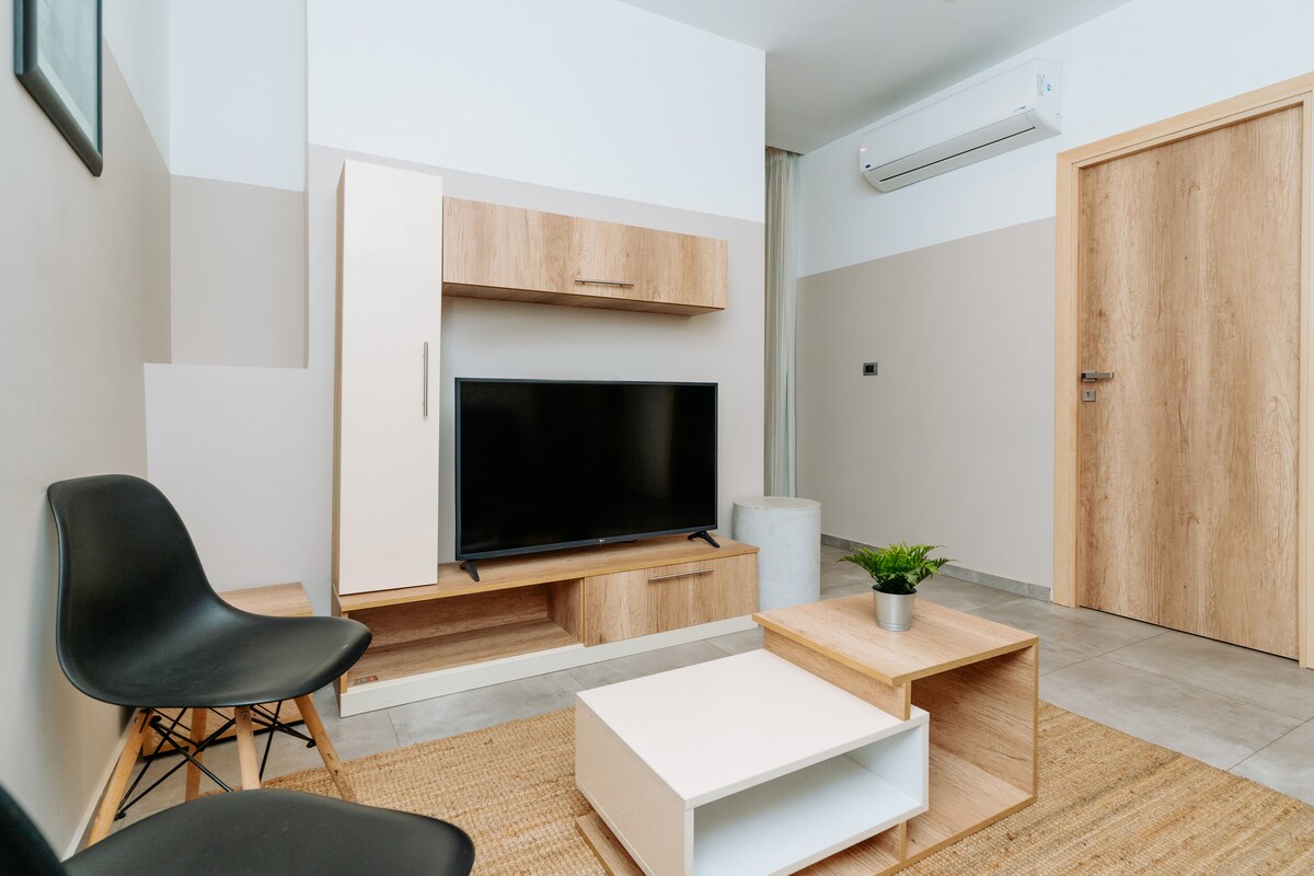 A modern living area is presented with a sleek television mounted on a wooden media console. A small coffee table sits on a textured area rug, accompanied by minimalist black chairs. Light enters through a nearby window, and a plant is positioned on the coffee table.