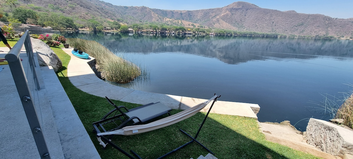 A tranquil lakeside area features a hammock positioned on a grassy bank, with a pathway leading to the water. Kayaks rest on the shore, surrounded by lush vegetation. The calm lake reflects the landscape, creating a serene view framed by gentle hills.