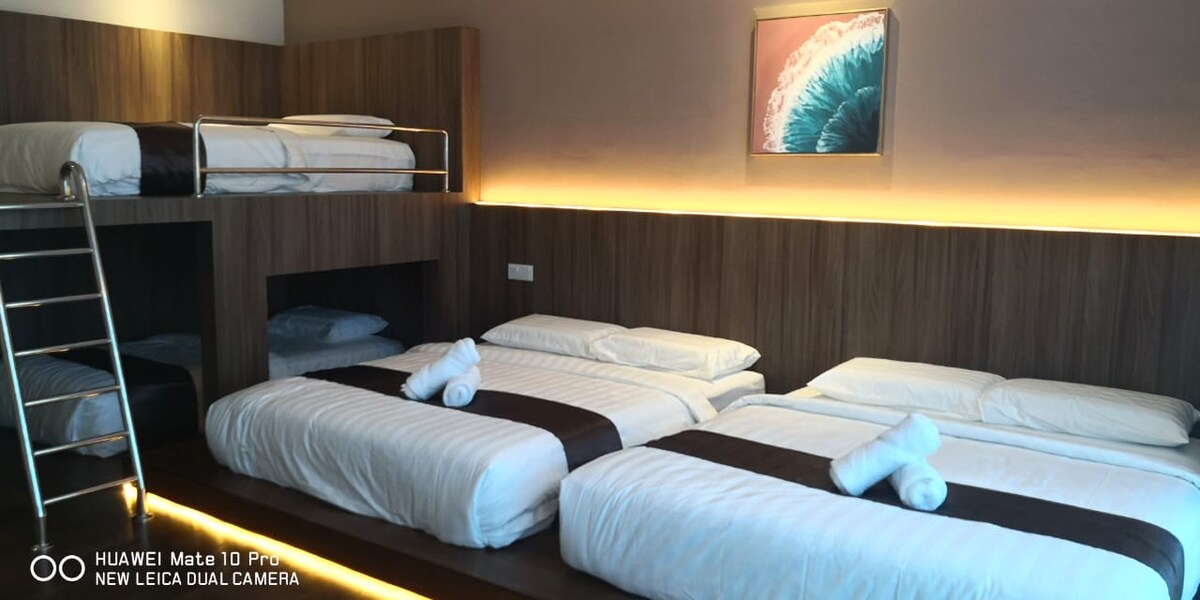 A modern sleeping area is showcased with multiple bedding arrangements. Two spacious beds are accompanied by a lofted bunk bed, featuring a sleek ladder. Soft linens are neatly arranged, and a decorative wall art piece adds a subtle touch to the warm wooden accents.