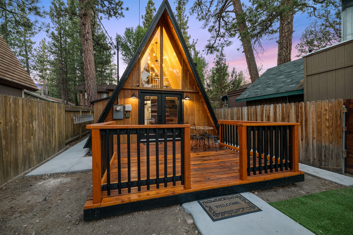 The exterior of the A-frame cabin features a wooden deck with black railings, providing an inviting entrance. Tall trees surround the property, contributing to a serene mountain ambiance. The cabin's large glass windows allow natural light to fill the interior while showcasing the cozy design.