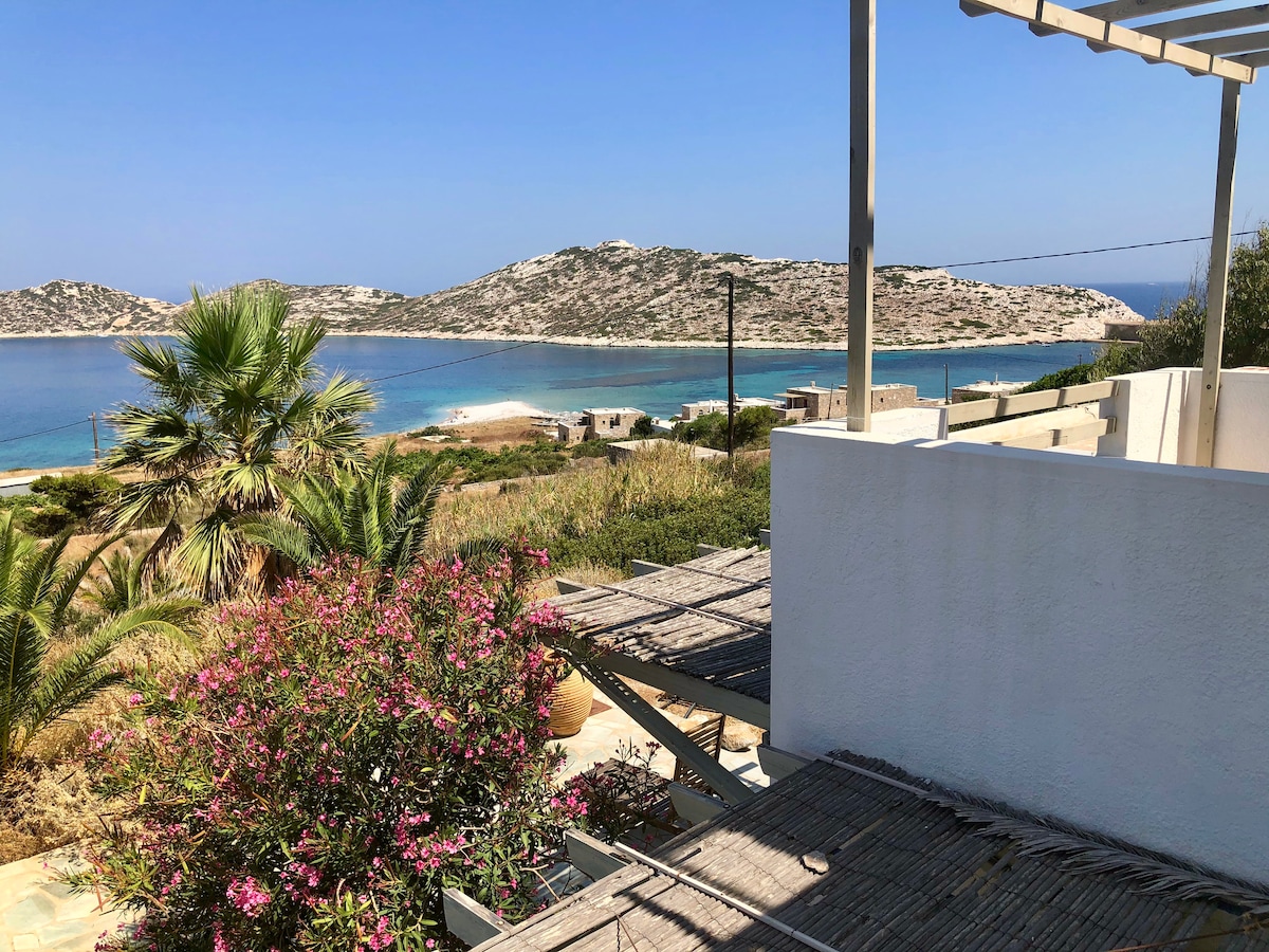 A spacious balcony offers a panoramic view of the sea and surrounding hills. Lush green palm trees and vibrant pink flowers are seen in the foreground. The clear blue waters reflect the sky, enhancing the serene setting.