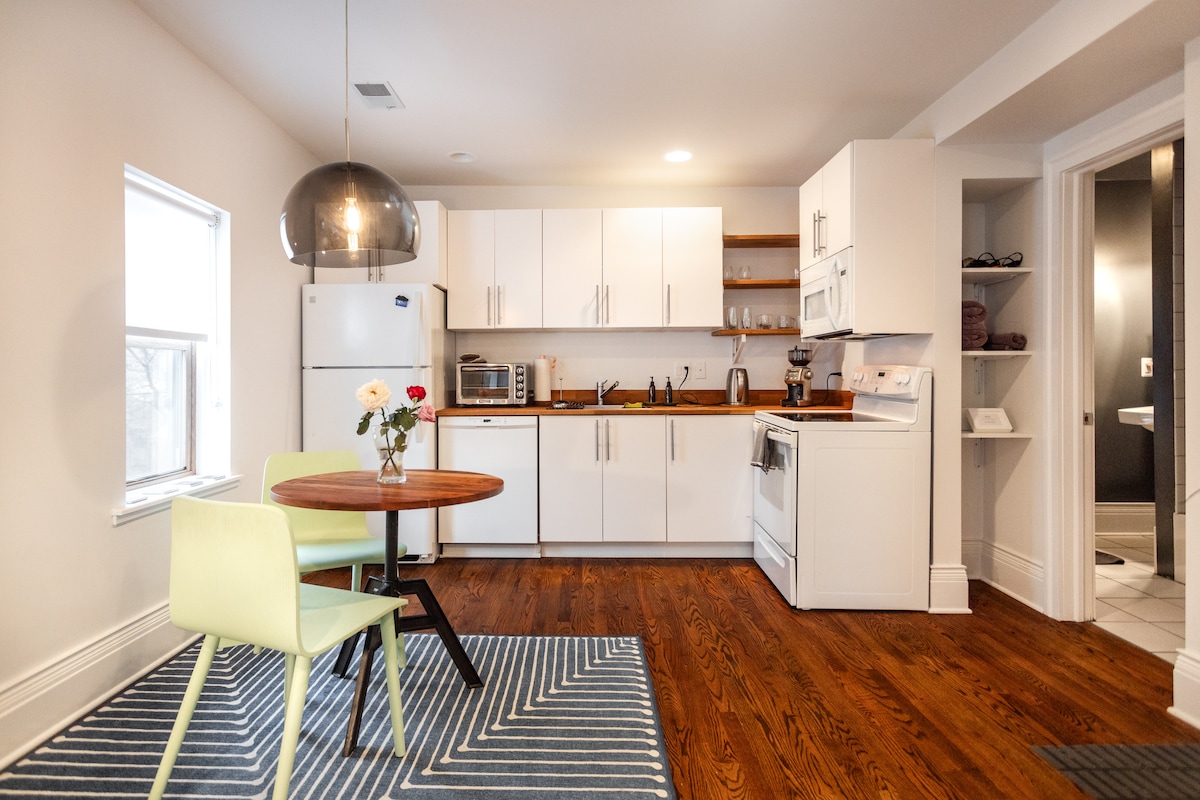 A modern kitchen is displayed with clean white cabinetry and a wooden countertop. A round dining table with two light green chairs sits on a navy blue and white striped rug. Natural light enters through the window, illuminating the space.