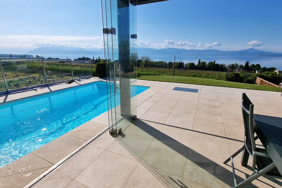 A private heated pool is visible against a backdrop of panoramic views of the lake and distant mountains. Spacious patio tiles lead from the pool to a large glass wall, allowing natural light to enhance the indoor-outdoor connection with the surrounding vineyards.