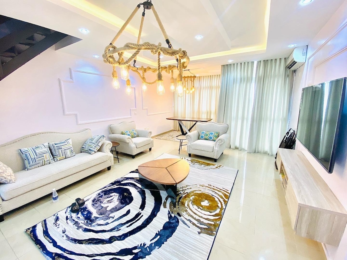 A light-filled living area features a comfortable seating arrangement with two sofas and decorative cushions. A large coffee table sits on a patterned rug, with a stylish chandelier overhead. The room is complemented by tall windows, allowing natural light to enhance the space.