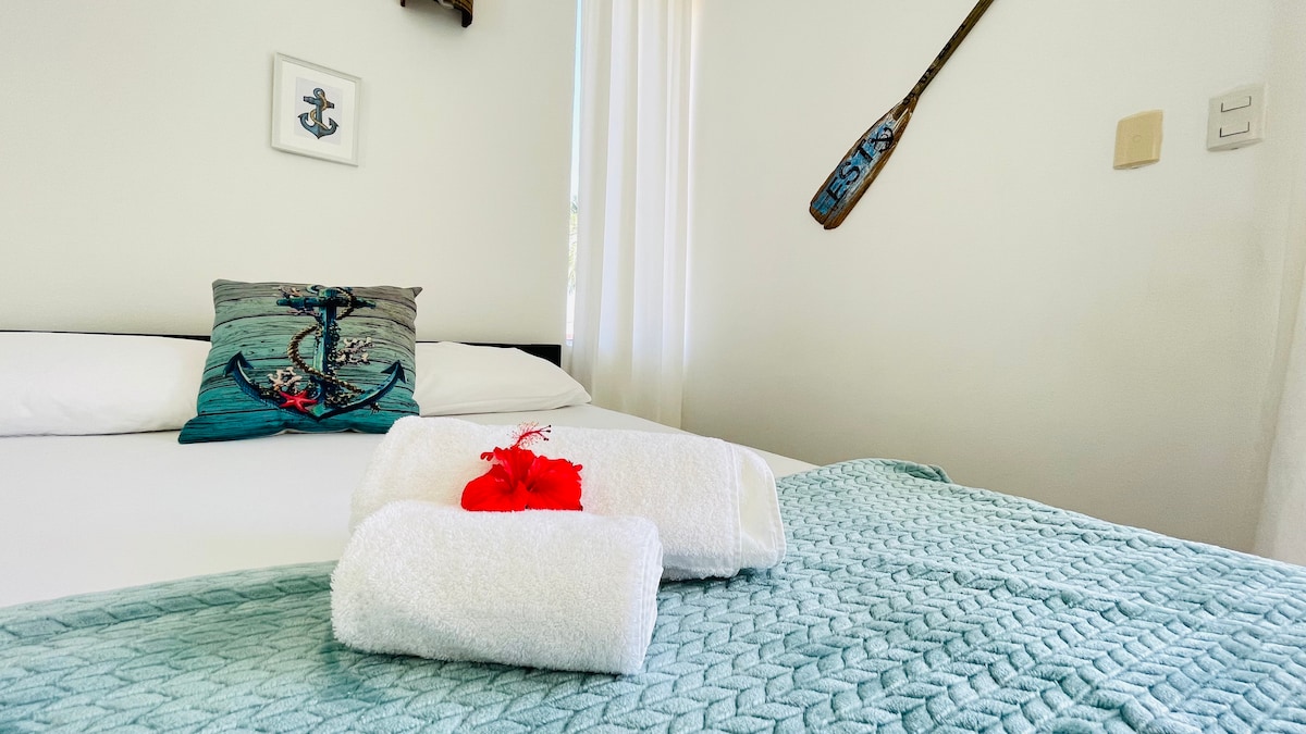 A cozy sleeping area features a spacious bed dressed in soft linens. Fresh towels are neatly arranged, accompanied by a decorative pillow featuring an anchor design. Light enters through sheer curtains, illuminating the room's serene white walls.