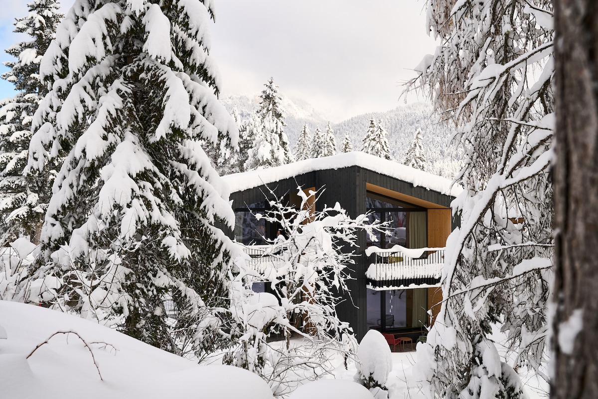 The modern structure is surrounded by a blanket of freshly fallen snow, with wooden accents contrasting against the dark exterior. Tall trees covered in snow frame the building, highlighting large glass windows that reveal glimpses of the interior.