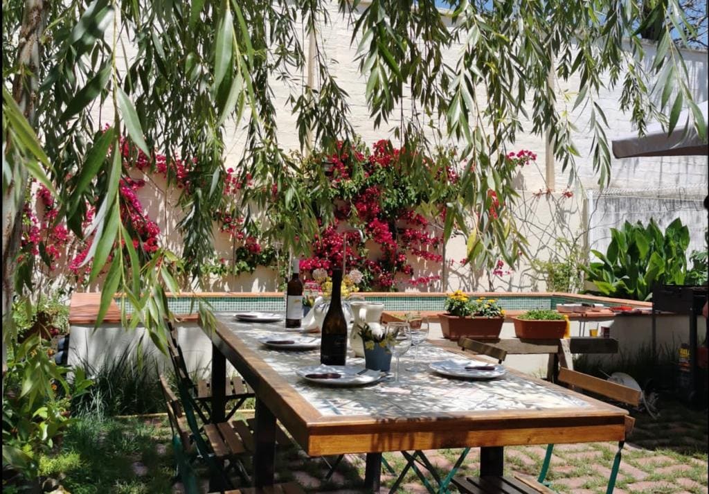 A shaded outdoor dining area features a large wooden table set with plates and bottles. Lush greenery and flowering plants create a vibrant backdrop, enhancing the inviting atmosphere for meals. The setting suggests a peaceful retreat surrounded by nature.