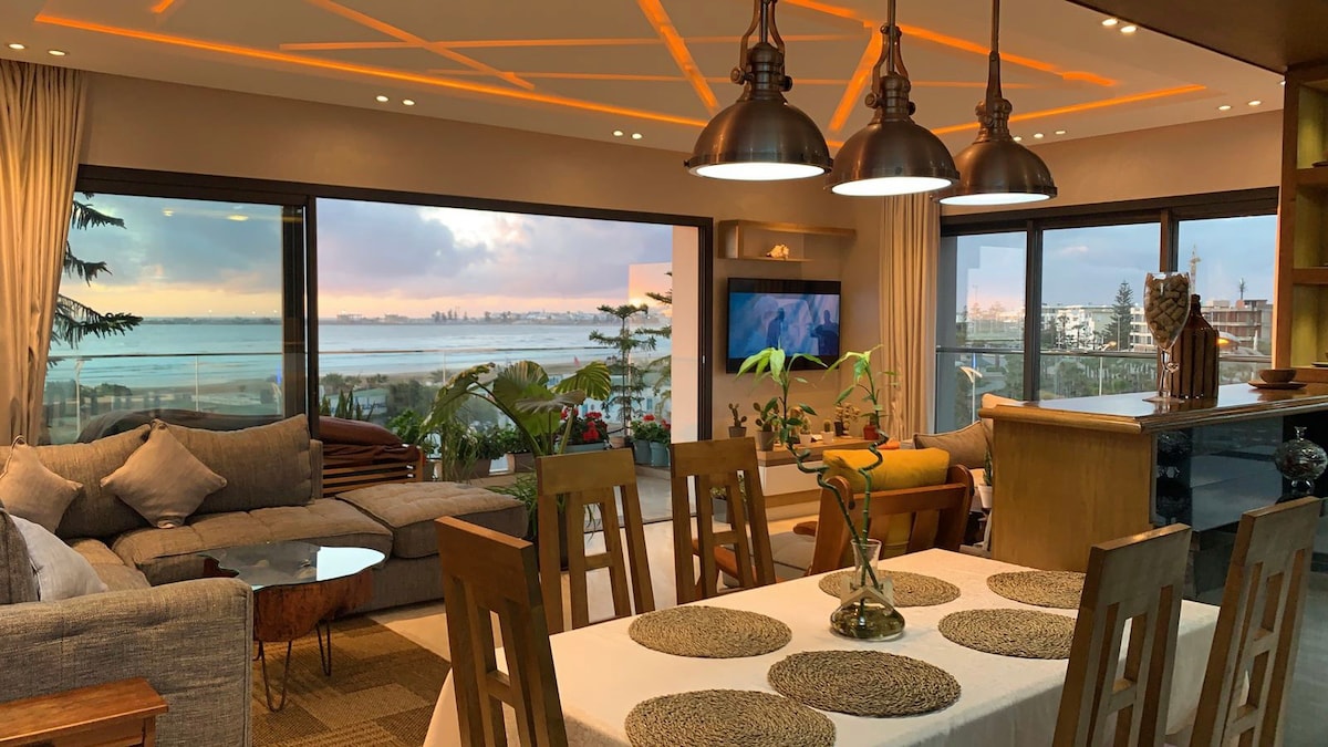 An open living space features large windows that offer views of the ocean and city skyline. A wooden dining table is set with decorative placemats, while a comfortable sofa area is enhanced by soft cushions. Warm lighting casts a welcoming glow throughout the room.