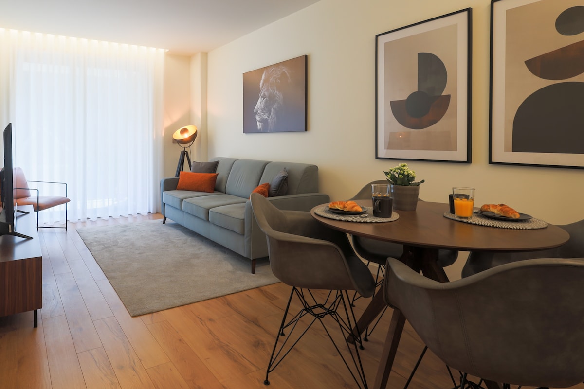 A comfortable living area is highlighted by a grey sofa and a round dining table set for four. Framed artwork adorns the walls, while natural light filters through sheer curtains, creating a welcoming ambiance. A small plant adds a touch of greenery to the space.