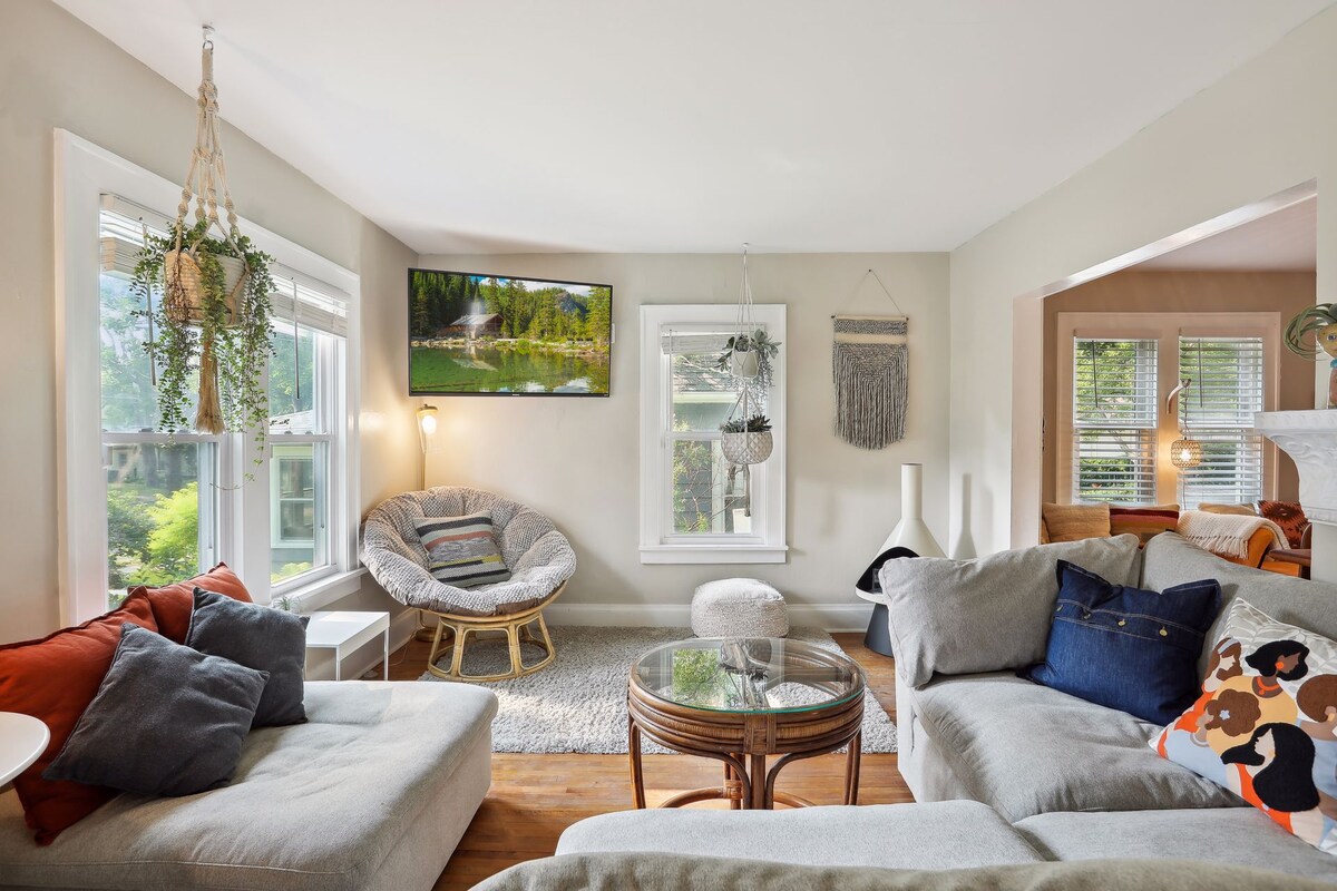 A light-filled lounge area showcases a comfortable sectional sofa and a round Papasan chair. A glass coffee table sits on a soft area rug, while decorative pillows and plants add a homey touch. Windows provide a view of the outdoors, enhancing the relaxing atmosphere.