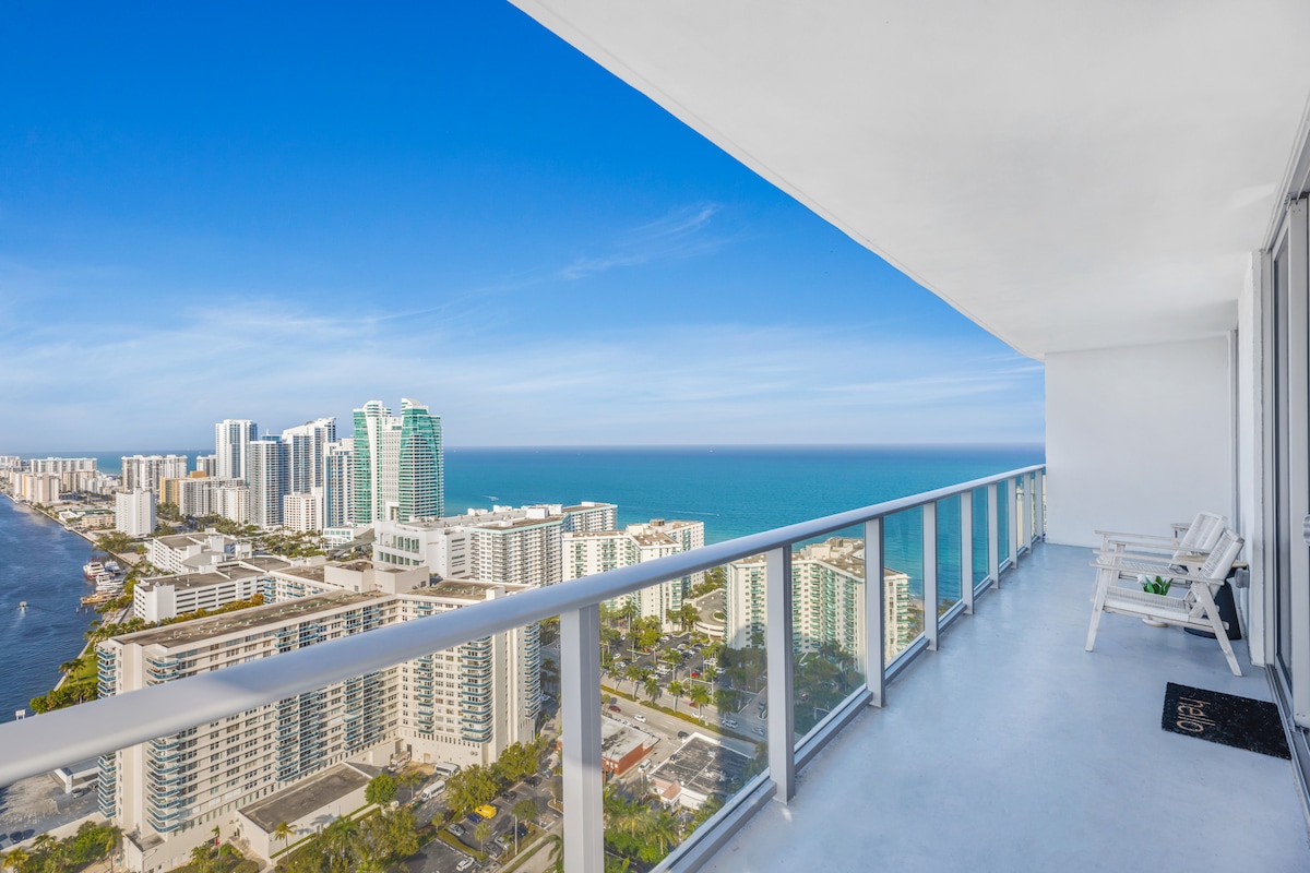 A spacious balcony overlooks stunning ocean views and city skyline, framed by glass railings. Two white lounge chairs provide a comfortable spot to take in the surroundings, while the clear blue sky enhances the vibrant coastal atmosphere.