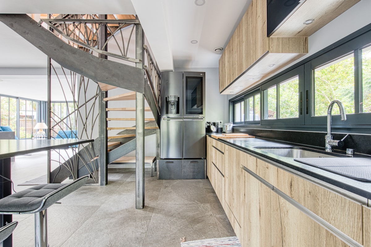 The kitchen features a sleek design with wooden cabinetry and black countertops. A stainless steel refrigerator is positioned alongside modern appliances. A spiral staircase leads to the upper level, enhancing the open concept layout. Large windows provide natural light and views of the surrounding greenery.