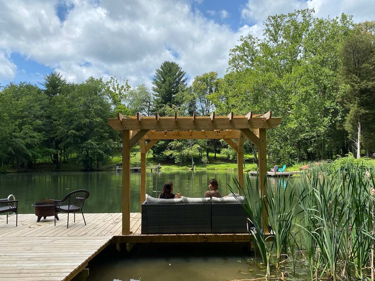 A shaded pergola sits on a wooden dock overlooking a serene lake, where two individuals are seen relaxing on a couch. Lush greenery surrounds the water, with chairs in the distance and a fire pit on the dock, inviting a tranquil experience.