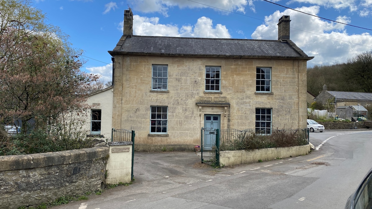 The house features a classic stone facade with large windows and a centered entrance door, framed by greenery. A gate leads to the front yard, while the surrounding area includes a quiet street and a glimpse of nearby buildings tucked among trees.
