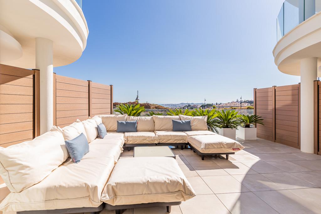 A spacious terrace features a large, comfortable seating area adorned with soft cushions in neutral tones, creating an open space for relaxation. Surrounding plants and wooden barriers provide a sense of privacy, complemented by a distant view of the landscape under clear blue skies.