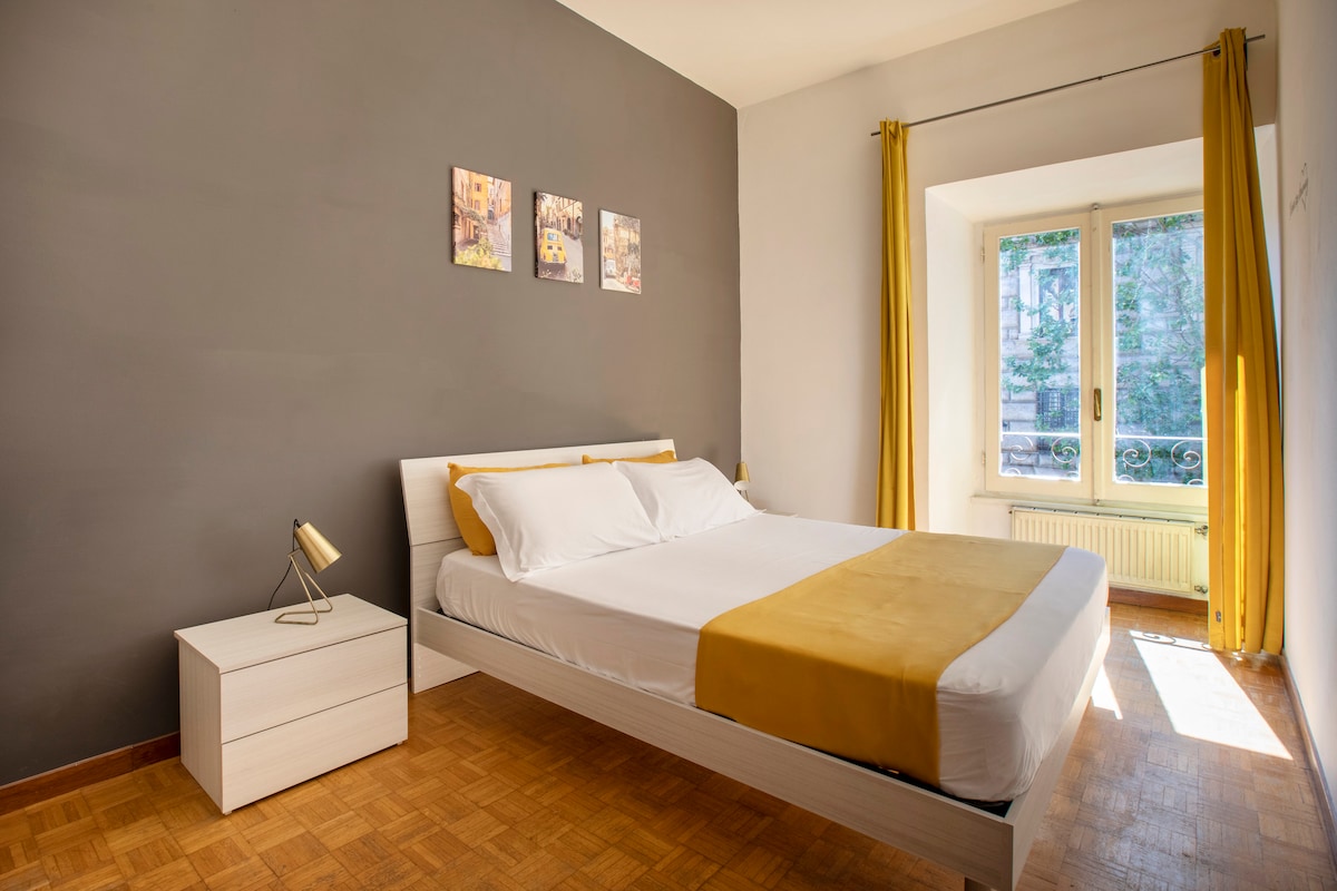 A bright and airy bedroom features a spacious bed topped with white linens and a yellow accent blanket. A minimalist nightstand with a lamp sits beside the bed. Natural light streams in through large windows adorned with yellow curtains, providing a view of the vibrant outdoor scenery.