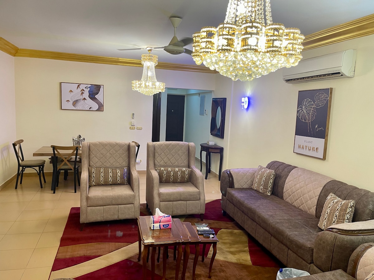 A spacious living area is furnished with two armchairs and a large sofa, arranged around a wooden coffee table. Elegant chandeliers provide ambient lighting, while a dining table is visible in the background, enhancing the overall functionality of the space.