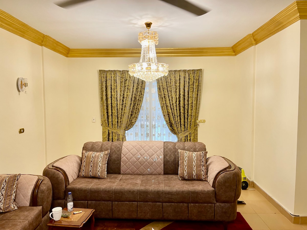A well-furnished living area is featured, showcasing a spacious sofa adorned with patterned throw pillows. Elegant curtains frame a large window, allowing natural light to brighten the pale walls. A decorative chandelier hangs from the ceiling, offering a touch of sophistication.