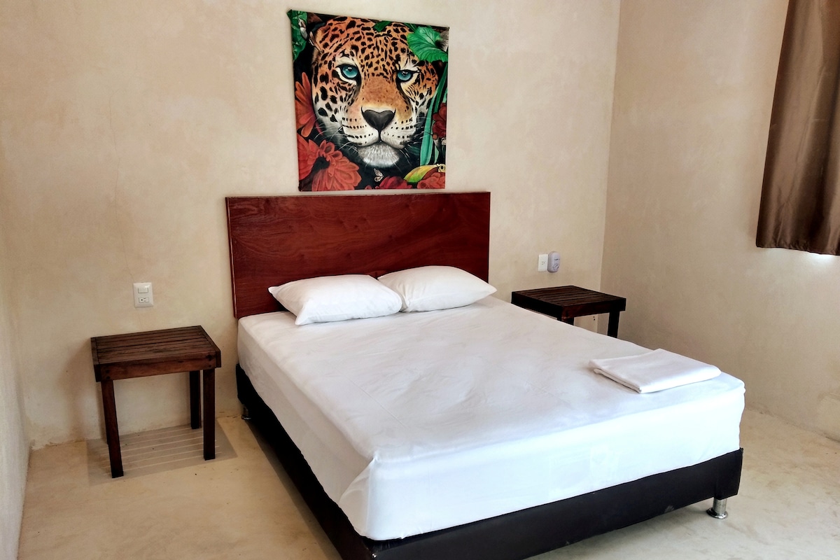 A cozy sleeping area features a spacious bed with neatly arranged white linens. Two wooden nightstands flank the bed, and a striking leopard artwork adorns the wall above the headboard. Natural light enters through a window, complemented by beige walls.
