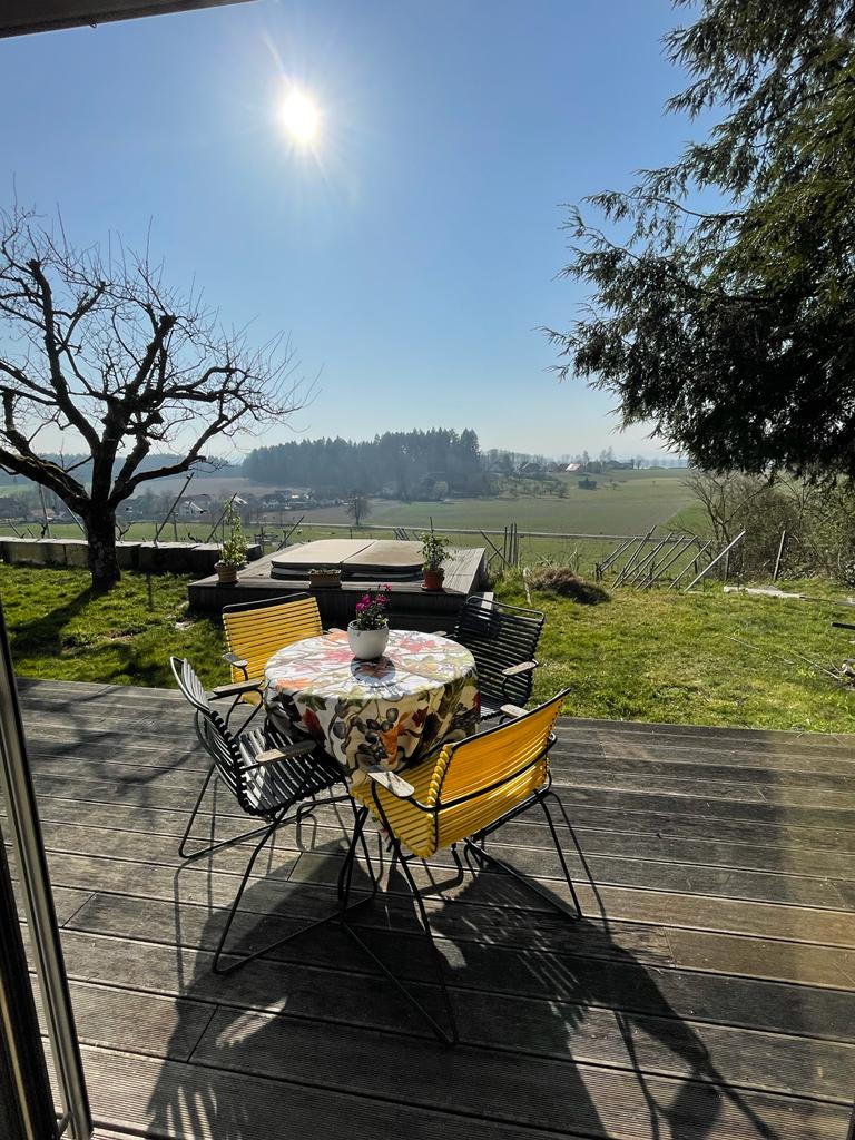 A terrace features a round table covered with a floral cloth and four chairs, set against a backdrop of green fields and distant hills. Sunlight shines brightly above, illuminating the peaceful outdoor space surrounded by greenery.