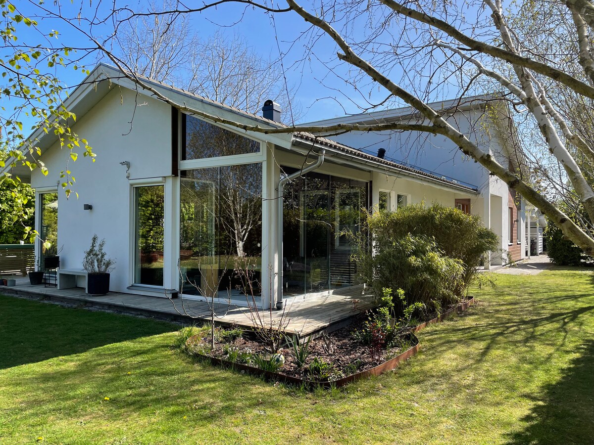 A modern house is set in a lush garden, featuring large glass windows and a spacious wooden deck. Surrounding greenery includes well-maintained shrubs and trees, contributing to an inviting outdoor environment. Sunlight highlights the clean lines of the architecture.
