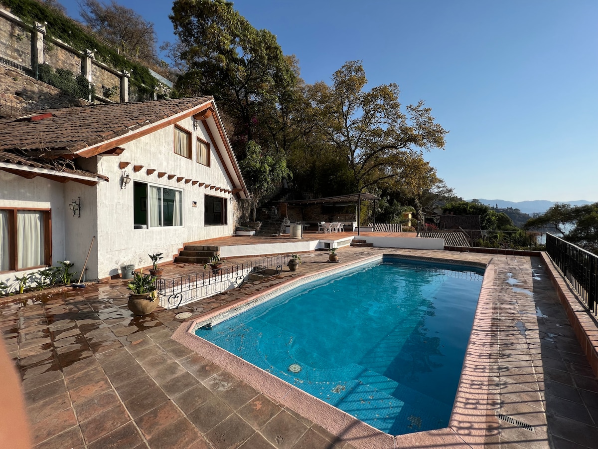 A spacious outdoor area features a rectangular swimming pool surrounded by stone tiles. Sun loungers are positioned nearby, and colorful potted plants add greenery. The house is visible with its white exterior, large windows, and a sloping roof, set against a backdrop of trees.