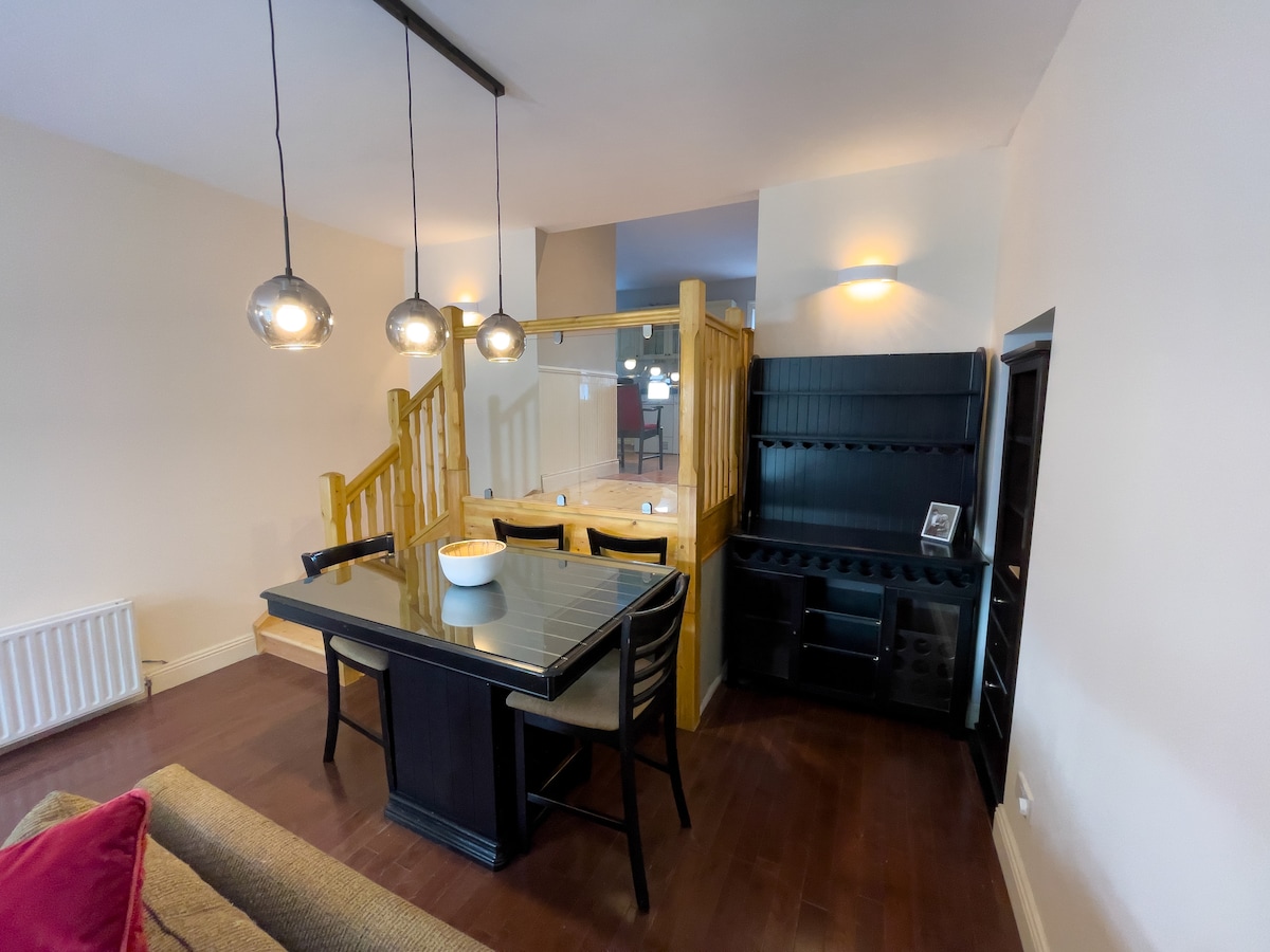 A dining area features a dark wooden table surrounded by black chairs, illuminated by pendant lighting above. The space is defined by a wooden partition, creating a sense of separation from the adjacent room. A stylish sideboard is positioned nearby, enhancing the functionality of the area.