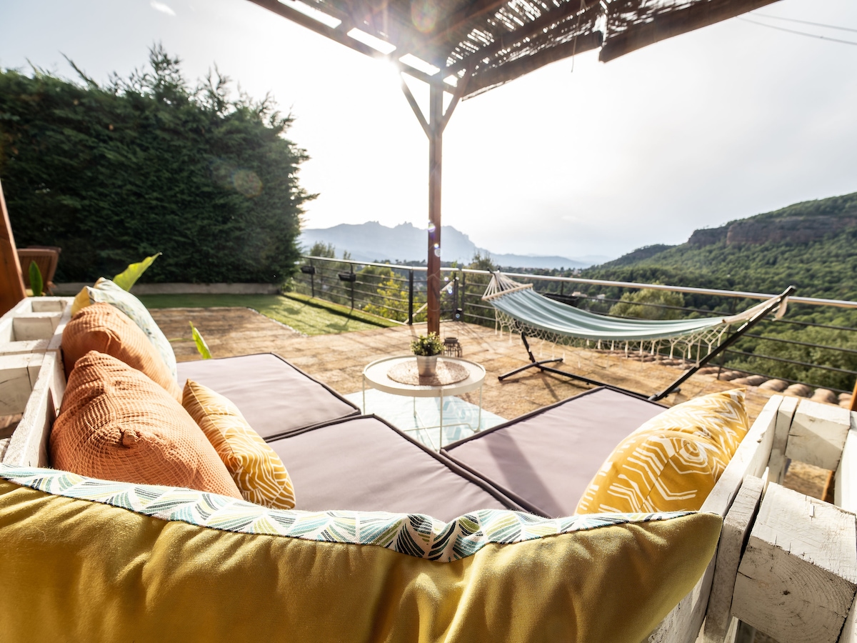 A cozy outdoor lounge area is visible, featuring plush cushions and a low table. A hammock is situated nearby, set against scenic views of the surrounding landscape. Lush greenery and distant mountains create a serene backdrop, enhancing the inviting atmosphere of the space.