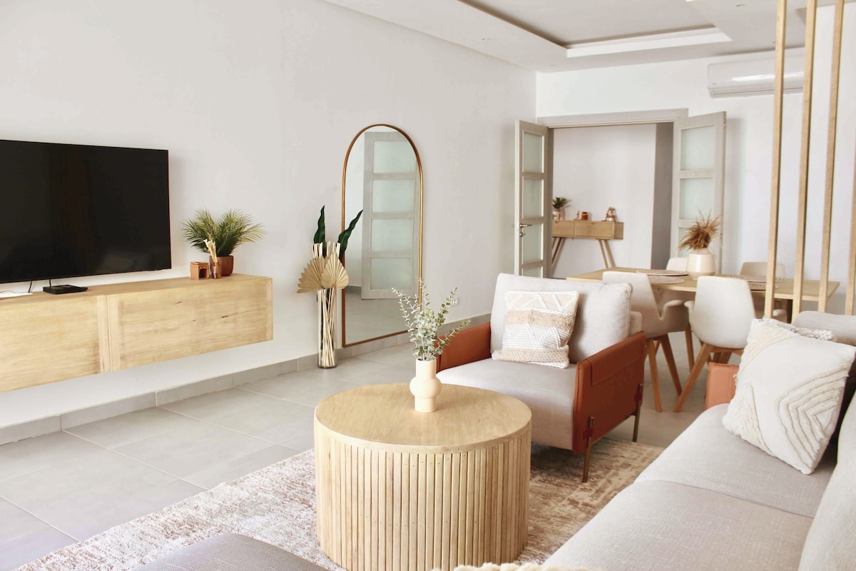 A bright and open living room features a comfortable sofa and a round coffee table. A flat screen TV is mounted on a wooden media console. Light enters through glass doors leading to a dining area with a small table and chairs, enhancing the inviting space.