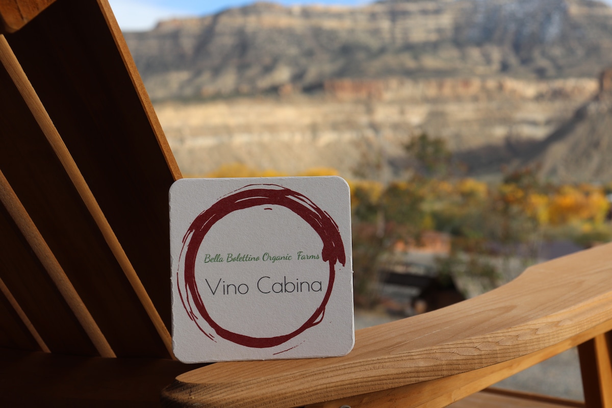 A coasters rests on a wooden chair, featuring the logo for Vino Cabina from Bella Bolettino Organic Farms. In the background, a vast landscape of layered rock formations and foliage is visible, indicative of the scenic surroundings.