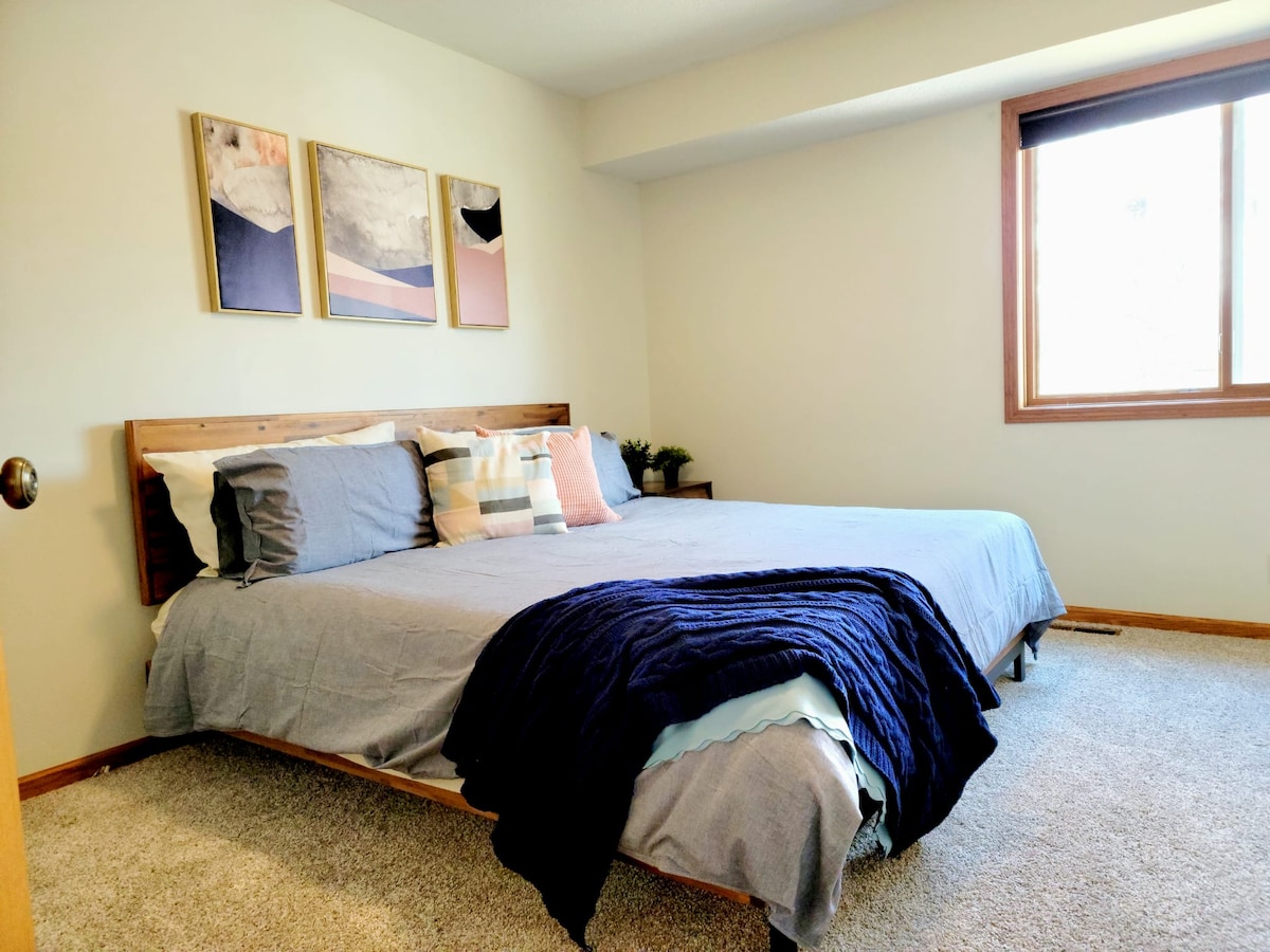 The master bedroom features a spacious bed dressed with layered bedding in calming tones. Three framed artworks hang above the bed, and natural light enters through a window adorned with simple blinds. Soft carpeting covers the floor, complementing the neutral wall color.