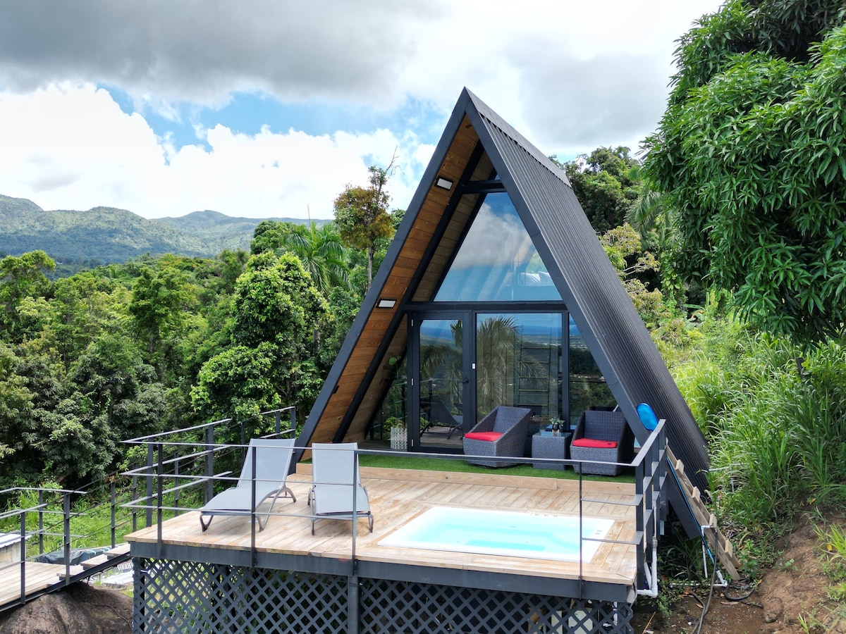 A modern A-frame structure is surrounded by lush greenery, featuring large glass windows that offer expansive mountain views. A wooden deck is equipped with two loungers and a small jacuzzi, inviting relaxation in a serene outdoor setting.