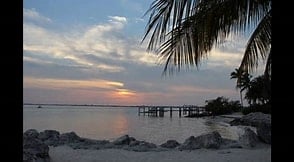 A serene view of the ocean at sunset is captured, with soft clouds reflecting warm tones across the sky. In the foreground, smooth rocks are framed by palm leaves, while a wooden dock extends into the calm waters, adding to the picturesque scenery.