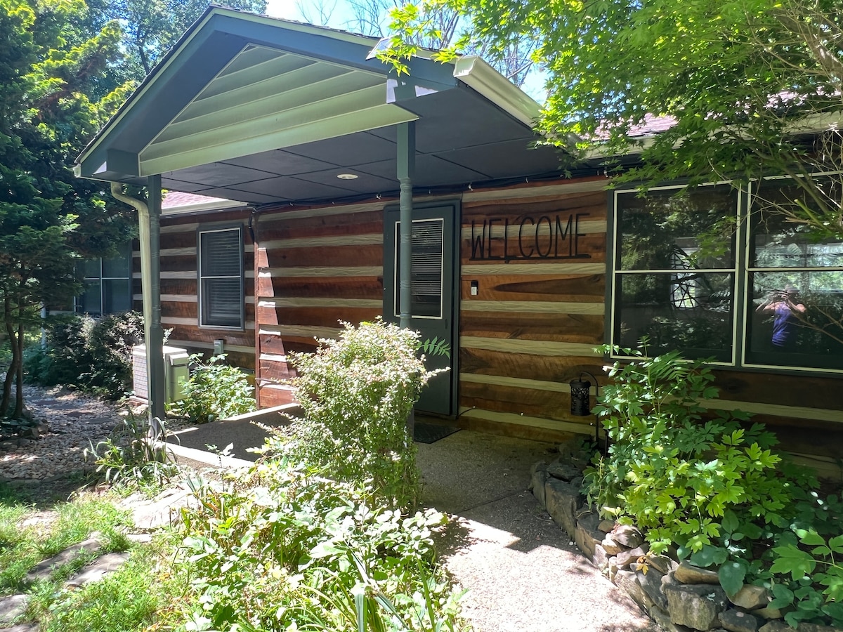 The cabin entrance features a rustic wooden façade accented by a welcoming sign. Lush greenery surrounds the pathway, leading to the front door, creating a natural and inviting atmosphere. Sunlight filters through the trees, highlighting the entrance's inviting design.